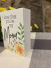 This is a 4" high wooden box sign that is painted white. It is one inch deep and with black lettering it says I love that you're my Mom. In the bottom right hand corner there is painted yellow flowers and green leaves.