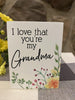 This is a 4" high wooden box sign that is painted white. It is one inch deep and with black lettering it says I love that you're my Grandma. In the bottom right hand corner there is painted yellow flowers and green leaves.