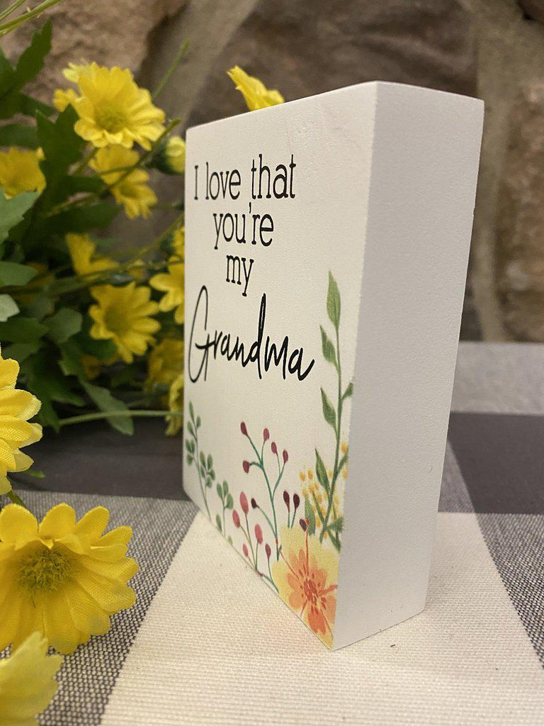 This is a 4" high wooden box sign that is painted white. It is one inch deep and with black lettering it says I love that you're my Grandma. In the bottom right hand corner there is painted yellow flowers and green leaves.