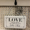 A farmhouse style white enamel sign with black lettering and hangs from a wire hanger. The sign says Love Grows Best in House like this.