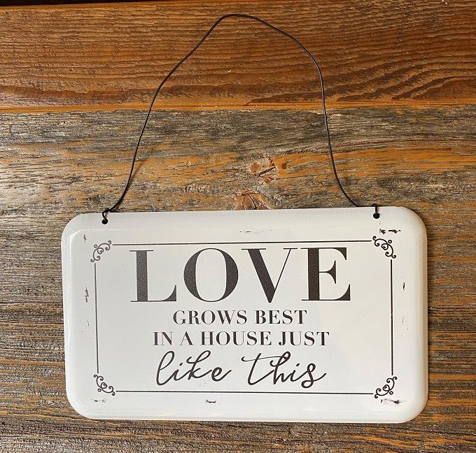 A farmhouse style white enamel sign with black lettering and hangs from a wire hanger. The sign says Love Grows Best in House like this.