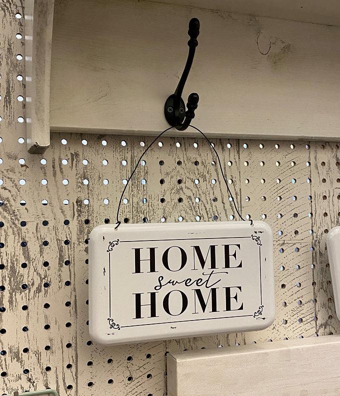 A white enamel sign with black lettering and hangs from a wire hanger. The sign says Home sweet home.