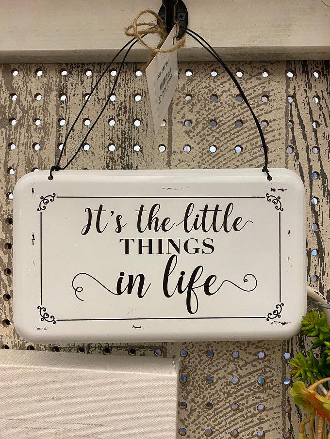 A farmhouse style white enamel sign with black lettering and hangs from a wire hanger. The sign says It's the Little Things in Life.