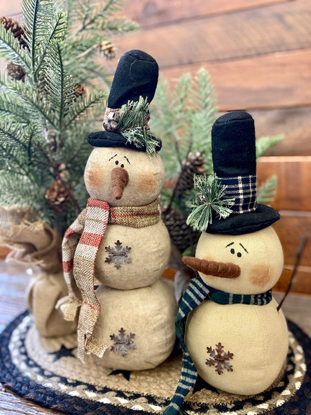 The Cranberrry plaid scarf snowman is a three stack snowman and has a black hat with cranberry and tan plaid trim and matching scarf. There  is a jingle bell trim on his hat and one metal snowlake on his middle and bottom stack. He has two twiggy arms. Also shown is the Green plaid scarf snowman. He has a black hat with jingle bell trims and two metal snowflakes. 