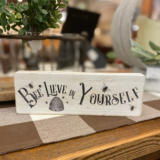 A wooden white washed block sign that has a icture of a bee hive and tiny honeybees ainted on it along with the words Bee Lieve in yourself.