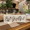 A whitewashed wooden block sign that will sit on a shelf. It says Bee Grateful and has a small painted honeybee.