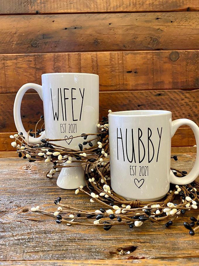 White ceramic mugs with the words WIFEY Est 2021 on one and HUBBY Est. 2021 on the other. Both have a small heart is printed in black under the lettering. The mug is customizable to the year.