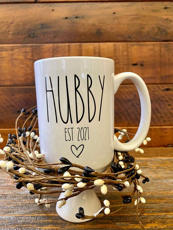 A white ceramic mug with the words HUBBY Est 2021 and a small heart is printed in black. The mug is customizable to the year.