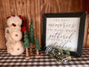 A square framed sign, with a black frame with some distressing on it. the sign is written in upper case and cursive style lettering and says: The best memories are made when gathered around the table. The sign is shown in a christmas theme setting.