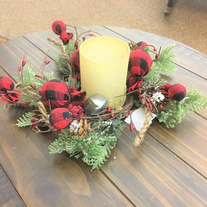 A beautiful Christmas wreath filled with snowy Christmas greens, silver jingle bells and round red and black check fabric coloured balls and pine cones, and sprigs of jute. Shown as a candle ring with a white candle in the center.