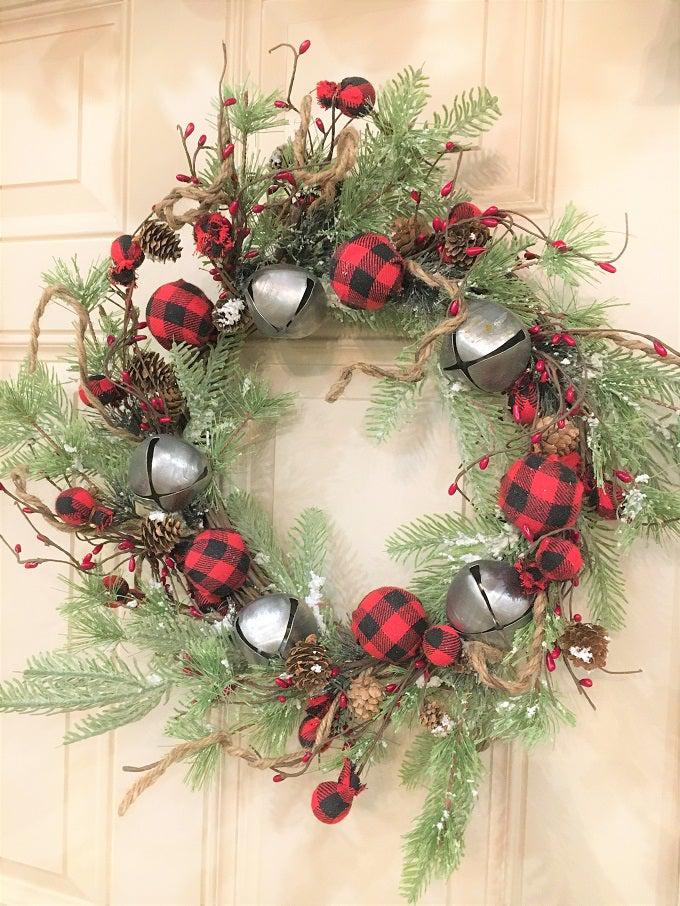 A beautiful Christmas wreath filled with snowy Christmas greens, silver jingle bells and round red and black check fabric coloured balls and pine cones and sprigs of jute rope.