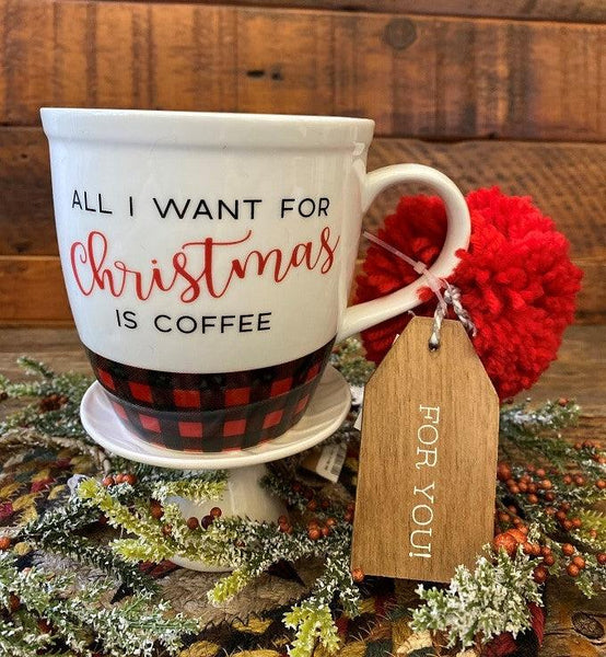 A white ceramic mug with a red pom pom and wooden gift tag ready for gifting. The mug has red buffalo plaid around the base and the wording All I want for Christmas is coffee.