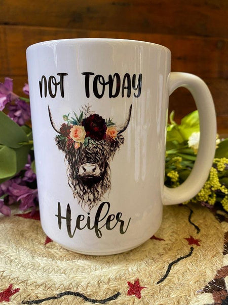 a white ceramic coffee mug that is imprinted on both sides with a pretty picture of a highland cow with flowers  on its head.The words "Not today Heifer" is written in a pretty script around the cow  image.