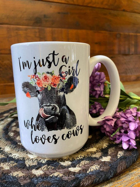 a white ceramic coffee mug that is imprinted on both sides with a  image of a black cow with pink flowers on its head. The words "I'm just a girl who loves cows" is written in a pretty script around the cow image.