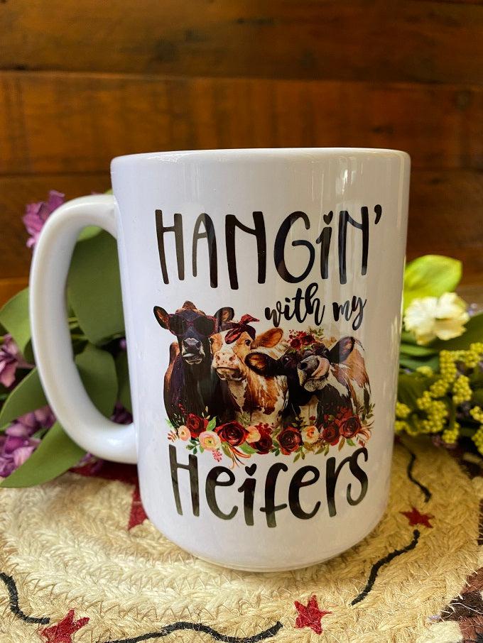 White ceramic mug imprinted on both sides with the words Hangin' with my Heifers and three brown heifer cows are pictured with a row of flowers in front of them. The cows are painted in a water-colour style.