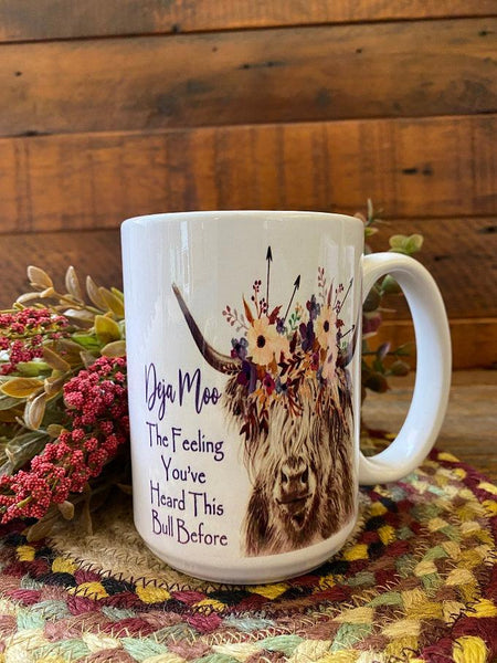 A white ceramic coffee mug that it is imprinted on both sides with a highland bull with flowers in its hair The saying is printed in black and says Deja Moo - The feeling that I've heard this bull before.