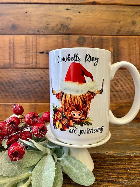 A white ceramic mug that says Cowbells Ring are you listening and has a picture of a highland cow with a santa hat.