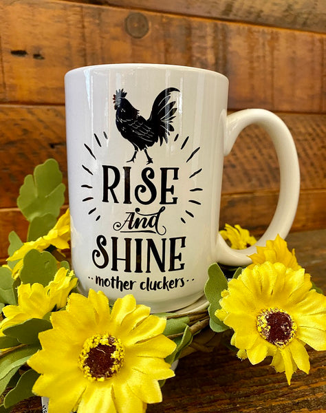 A white ceramic mug with a black rooster that says Rise and shine mother cluckers.